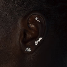Load image into Gallery viewer, A close-up side profile of a person's ear wearing multiple small silver ear cuffs, including one with a floral design and one with script lettering, against a dark background.
