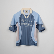 Load image into Gallery viewer, A vintage-style short-sleeve soccer jersey from Curacao, featuring a light blue body, darker blue sleeves and sides, a beige collar, and light pink side panels. The jersey has 'CURAÇÃO' printed across the chest with 'The Islands' below it, and various sponsor logos.
